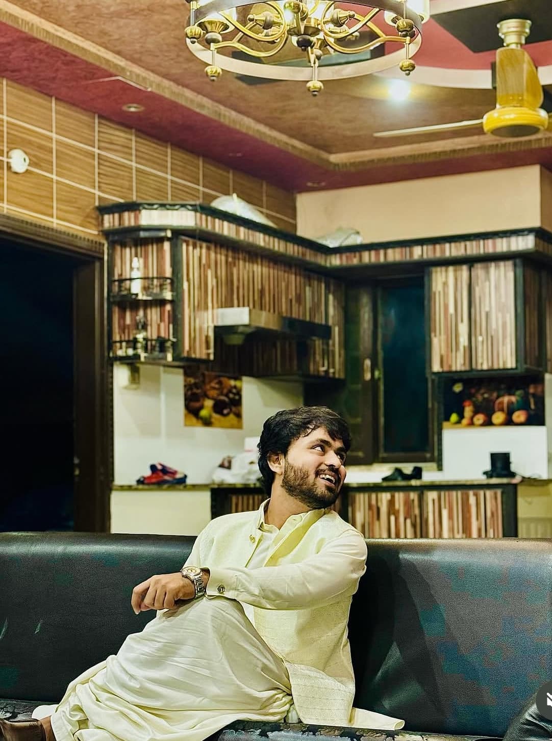 Smiling bearded man in a cream traditional outfit sits on a couch, looking away.